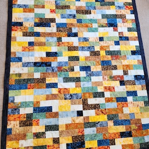May include: A rectangular quilt with a patchwork design. The quilt features a variety of rectangular fabric pieces in shades of yellow, blue, orange, brown, and white. The quilt is bordered with a dark blue fabric.