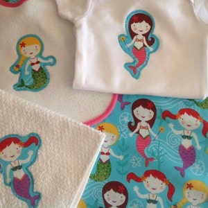 May include: A set of three white fabric items with embroidered mermaid designs. The items include a bib, a burp cloth, and a onesie. The mermaids are all wearing pink and purple swimsuits and have long, flowing hair. The background of the fabric is a light blue with white waves and seashells.