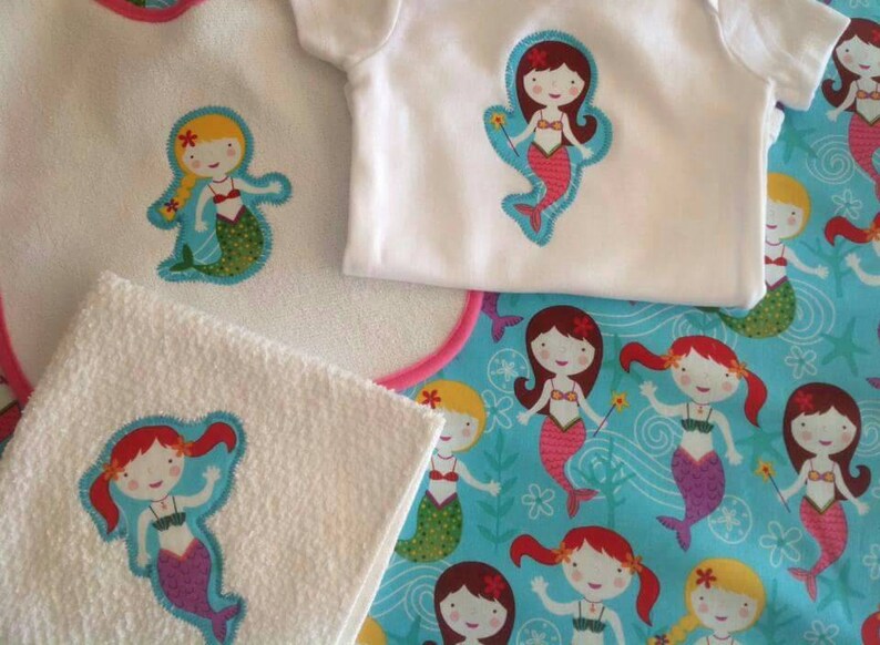 May include: A set of three white fabric items with embroidered mermaid designs. The items include a bib, a burp cloth, and a onesie. The mermaids are all wearing pink and purple swimsuits and have long, flowing hair. The background of the fabric is a light blue with white waves and seashells.