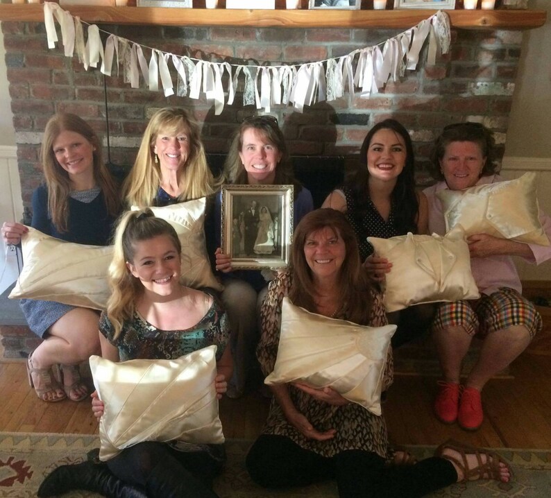 Puede incluir: Un grupo de mujeres reunidas alrededor de una chimenea, sosteniendo almohadas de sat&eacute;n blanco. Est&aacute;n celebrando una boda con una foto enmarcada de una novia y un novio.