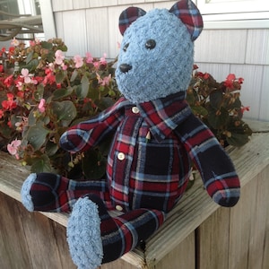 May include: A handmade teddy bear with a blue head and feet, and a plaid shirt and pants in red, navy, and light blue. The bear is sitting on a wooden surface near a flower box with pink flowers.