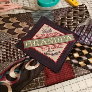 Puede incluir: Una almohada de tela azul oscuro con un dise&ntilde;o de patchwork hecho de diferentes corbatas estampadas. El centro de la almohada presenta un parche en forma de diamante con un gr&aacute;fico de autom&oacute;vil vintage y el texto "NOT JUST AN OLD MODEL GRANDPA BUT A CLASSIC".
