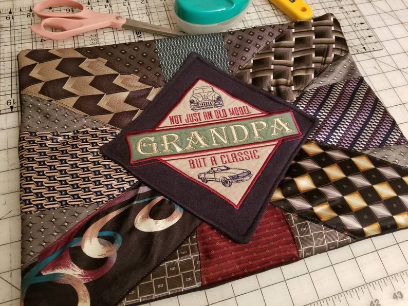 Puede incluir: Una almohada de tela azul oscuro con un dise&ntilde;o de patchwork hecho de diferentes corbatas estampadas. El centro de la almohada presenta un parche en forma de diamante con un gr&aacute;fico de autom&oacute;vil vintage y el texto "NOT JUST AN OLD MODEL GRANDPA BUT A CLASSIC".