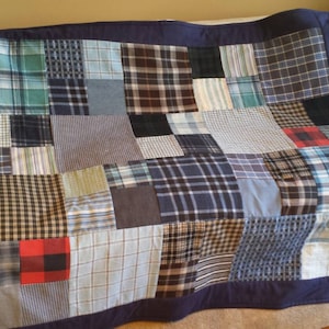May include: A patchwork quilt with a navy blue border, composed of various plaid and patterned fabrics in shades of blue, gray, and red. The quilt is draped over a cream-colored sofa, showcasing its design and texture.