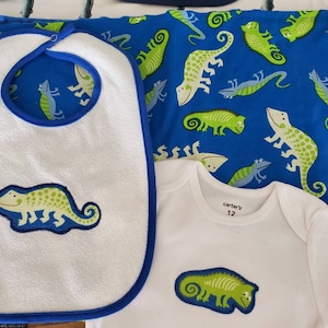May include: A white bib with a blue trim and a green chameleon design. A white onesie with a blue trim and a green chameleon design. The onesie has a tag that says "carter's" and "12".
