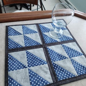 May include: A square quilted coaster with a geometric pattern of blue polka dots and gray triangles. The coaster has a dark gray border and is shown with a wine glass on top. The coaster is on a light-colored surface.