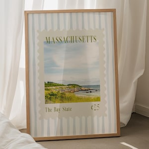 Op de afbeelding: Een ingelijste print met een kustlandschap uit Massachusetts, met de tekst "MASSACHUSETTS" en "The Bay State". De print heeft een lichtblauwe en witte gestreepte achtergrond en een houten lijst. De print wordt weergegeven in de buurt van een bed en een kleine houten tafel.