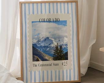 May include: A framed print featuring a Colorado landscape, resembling a postage stamp design. The print showcases a mountain range under a cloudy sky, with the text "COLORADO" and "The Centennial State". The frame has a light wood finish and is set against a blue and white striped background.