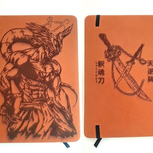May include: A tan notebook with a dark brown laser-engraved illustration of a muscular humanoid figure on the front cover and a pair of crossed swords with chains and Japanese text on the back cover, featuring a black elastic closure band.