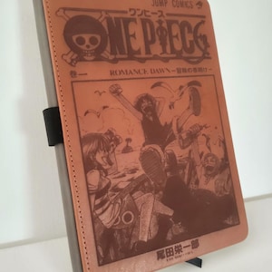 May include: A brown leather journal with the "One Piece" manga cover art. The cover shows the title, "ONE PIECE", and Japanese text, along with character illustrations. The journal has a grey spine and a black elastic band closure.