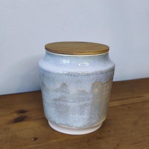 Puede incluir: Un tarro de cerámica con un esmalte azul claro y gris, coronado con una tapa de madera. El tarro tiene un cuerpo ancho y una base más estrecha. La tapa es de color madera natural. El tarro está sobre una superficie de madera.