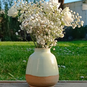Peut inclure: Un vase en céramique, vert clair en haut et marron en bas, contient un bouquet de fleurs blanches. L'arrangement comprend des roses blanches et de petites fleurs blanches délicates. Le vase est posé sur une surface en bois.