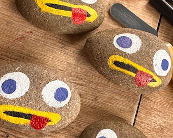 May include: Four painted rocks with faces. Each rock has a yellow mouth, white eyes, and blue pupils. The rocks have red tongues sticking out. 