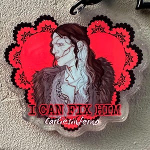 May include: A clear acrylic keychain featuring a portrait of a person with long dark hair and pale skin, set against a red heart-shaped background with a black floral border. The text "I CAN FIX HIM" and "carliesinferno" are printed below the portrait.