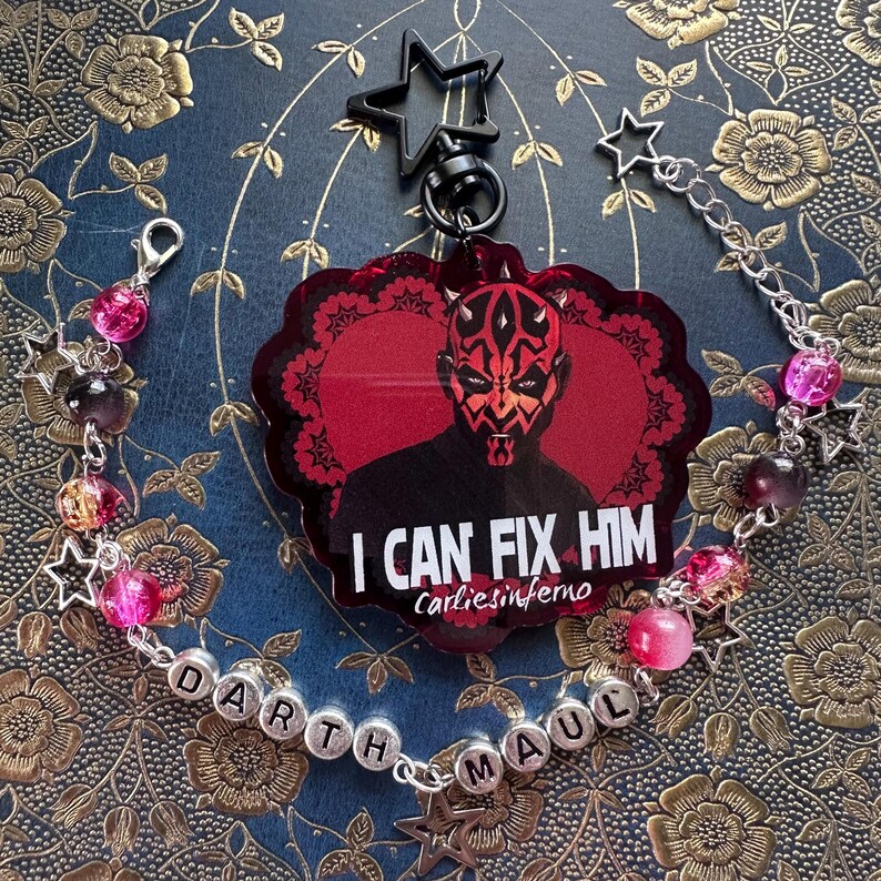 Może przedstawiać: Czerwono-czarny brelok Darth Maul z napisem "I CAN FIX HIM" i pasującą bransoletką. Bransoletka ma r&oacute;żowe i fioletowe koraliki, zawieszki w kształcie gwiazd i napis "Darth Maul".