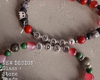 May include: Two beaded bracelets featuring red, green, and grey stone beads with silver-toned spacers. One bracelet includes a white ghost-shaped charm and silver letter beads spelling GHOST SOAP. The text NEW DESIGN Glass + Stone Beads Resin free is visible.