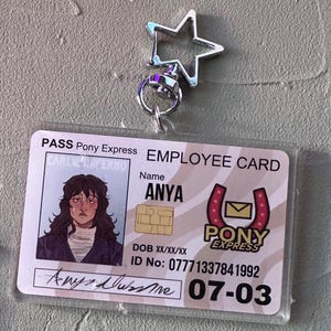 Peut inclure: Une carte d'identité d'employé en plastique avec une illustration de dessin animé d'une personne aux cheveux bruns et une chemise bleue. La carte porte le texte "PASS Pony Express EMPLOYEE CARD" et "PONY EXPRESS" avec un logo en forme de fer à cheval. La carte comprend également le nom "ANYA", un numéro d'identification et la date "07-03".