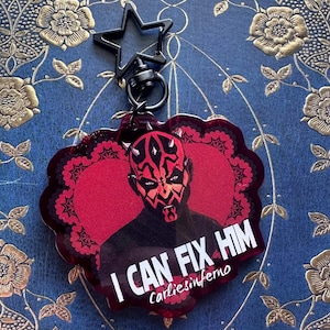 May include: A red and black keychain featuring a portrait of Darth Maul with the text "I CAN FIX HIM" and the name "carliesinferno". The keychain has a black star-shaped clasp.