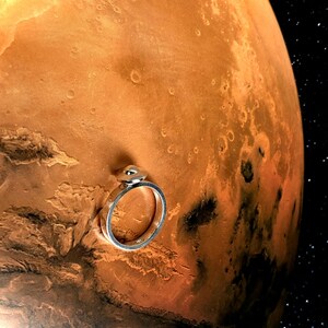 May include: A silver ring with a small, round, light-colored stone set on top. The ring is set against a backdrop of the planet Mars, with its reddish-brown surface and craters. The background includes a starry night sky.