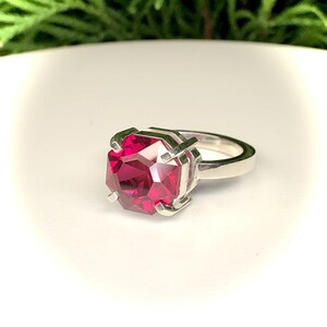 May include: A silver ring featuring a large, faceted, deep red gemstone. The gemstone is held in place by a silver setting with four prongs. The ring is set against a white background with green foliage in the background.