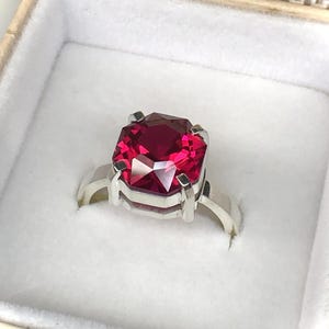 May include: A silver ring with a large, faceted, rectangular, deep red gemstone. The ring is set in a square-shaped setting and is presented in a white jewelry box with a gold-colored trim.