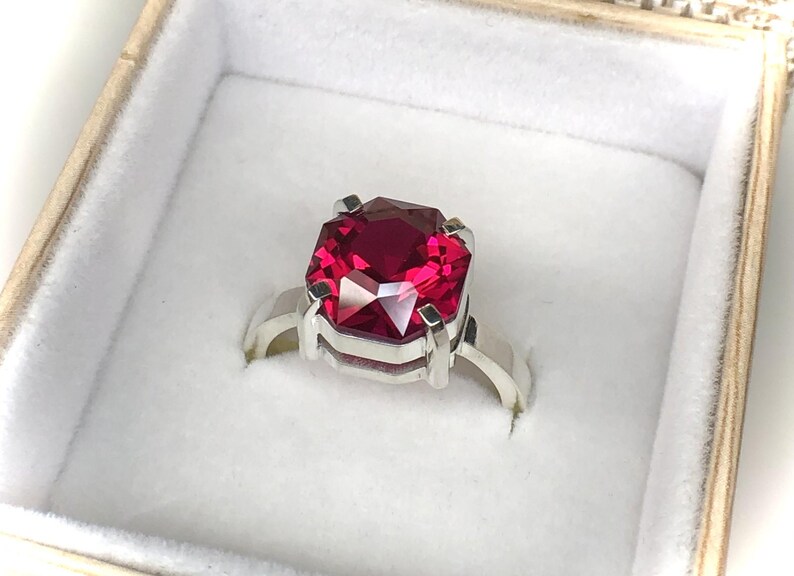 May include: A silver ring with a large, faceted, rectangular, deep red gemstone. The ring is set in a square-shaped setting and is presented in a white jewelry box with a gold-colored trim.