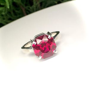 May include: A silver ring featuring a large, faceted, octagonal-cut, ruby-red gemstone. The gemstone is set with four prongs, and the band is a simple, polished silver. The ring is displayed on a white surface, with green foliage in the background.