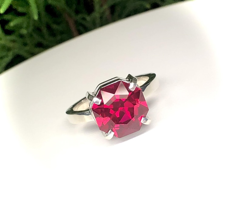 May include: A silver ring featuring a large, faceted, octagonal-cut, ruby-red gemstone. The gemstone is set with four prongs, and the band is a simple, polished silver. The ring is displayed on a white surface, with green foliage in the background.