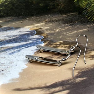 May include: A pair of silver surfboard earrings. Each earring features a small, detailed surfboard charm with a dark gray center. The earrings have a simple hook design, perfect for everyday wear. The background shows a beach scene.