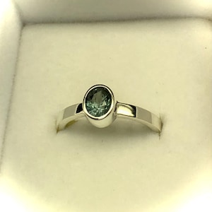 May include: A silver-toned ring featuring a single oval-cut, blue-green gemstone set in a smooth bezel setting. The band is polished and solid, with a clean, reflective surface that highlights the central stone's multifaceted, deep teal appearance.