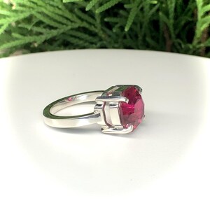 May include: A silver ring featuring a large, faceted, ruby-red gemstone. The ring has a simple band and a square setting that holds the stone securely. The gemstone is the focal point, catching the light and displaying its vibrant color.
