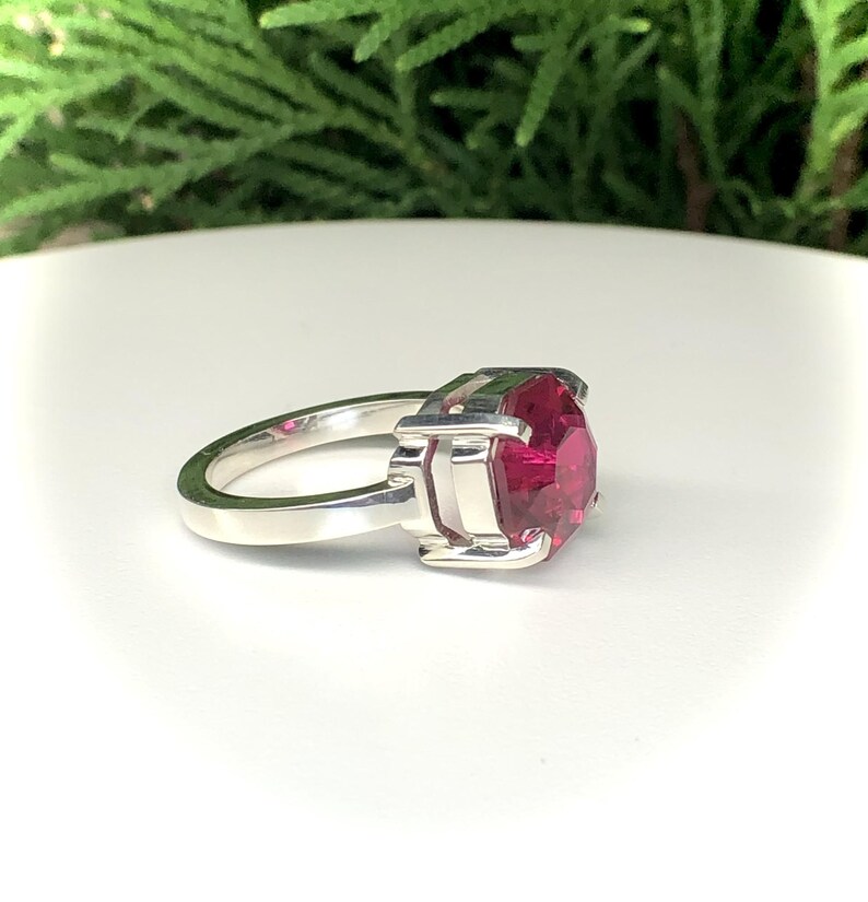 May include: A silver ring featuring a large, faceted, ruby-red gemstone. The ring has a simple band and a square setting that holds the stone securely. The gemstone is the focal point, catching the light and displaying its vibrant color.