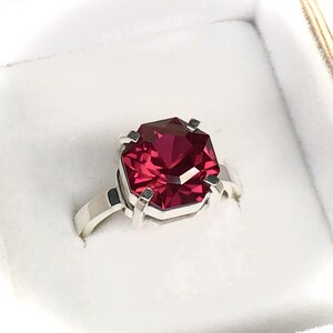 May include: A silver ring featuring a large, faceted, rectangular-cut, deep red gemstone. The gemstone is set in a four-prong setting, and the band is a simple, polished silver. The ring is displayed in a white jewelry box.
