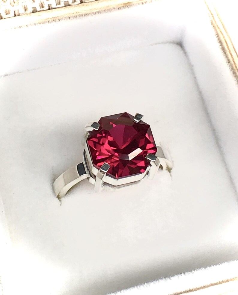 May include: A silver ring featuring a large, faceted, rectangular-cut, deep red gemstone. The gemstone is set in a four-prong setting, and the band is a simple, polished silver. The ring is displayed in a white jewelry box.