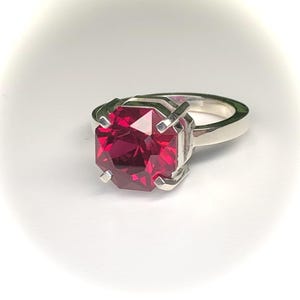 May include: A silver ring featuring a large, faceted, rectangular-cut, deep red gemstone. The gemstone is set in a four-prong setting, and the band is a simple, polished silver. The ring is displayed on a white background.