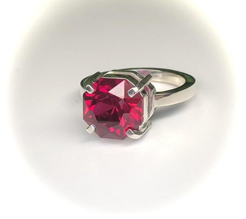 May include: A silver ring featuring a large, faceted, rectangular-cut, deep red gemstone. The gemstone is set in a four-prong setting, and the band is a simple, polished silver. The ring is displayed on a white background.