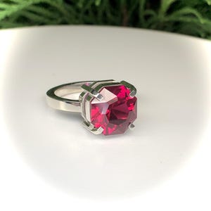 May include: A silver ring featuring a large, faceted, square-cut, ruby-red gemstone. The gemstone is held in place by four silver prongs. The ring band is a simple, polished silver.
