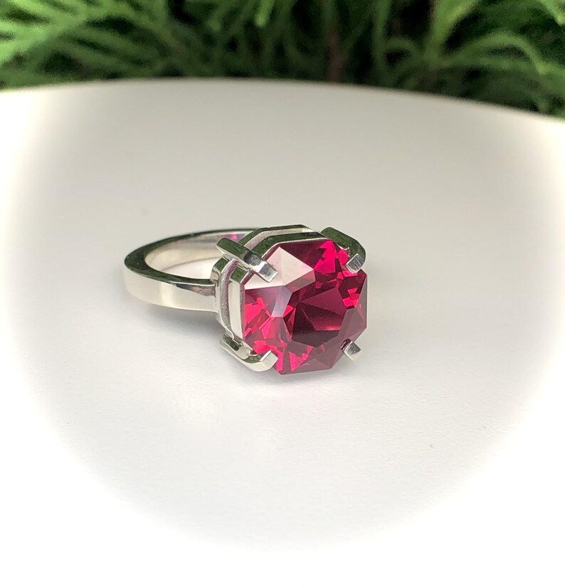 May include: A silver ring featuring a large, faceted, square-cut, ruby-red gemstone. The gemstone is held in place by four silver prongs. The ring band is a simple, polished silver.