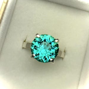 May include: A silver ring with a large, round, faceted teal gemstone. The gemstone is the focal point, set with four prongs. The ring is presented in a white jewelry box, with a gold-colored trim.