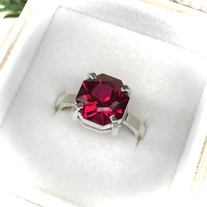 May include: A silver ring featuring a large, faceted, rectangular-cut, deep red gemstone. The gemstone is set in a four-prong setting, and the band is a simple, polished silver. The ring is presented in a white jewelry box.