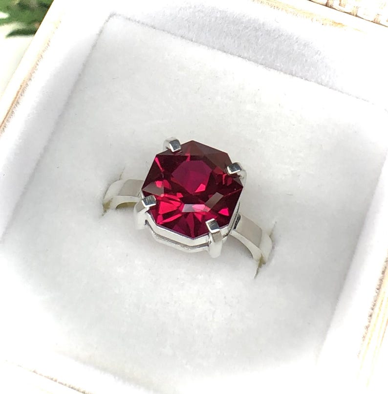May include: A silver ring featuring a large, faceted, rectangular-cut, deep red gemstone. The gemstone is set in a four-prong setting, and the band is a simple, polished silver. The ring is presented in a white jewelry box.