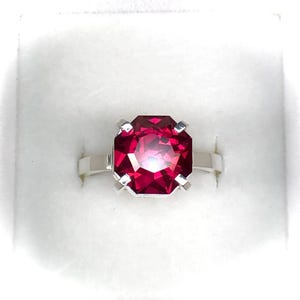 May include: A silver ring featuring a large, faceted, rectangular-cut ruby gemstone. The ruby is a vibrant shade of red, and the ring is set in a simple, elegant design. The ring is displayed in a white box.