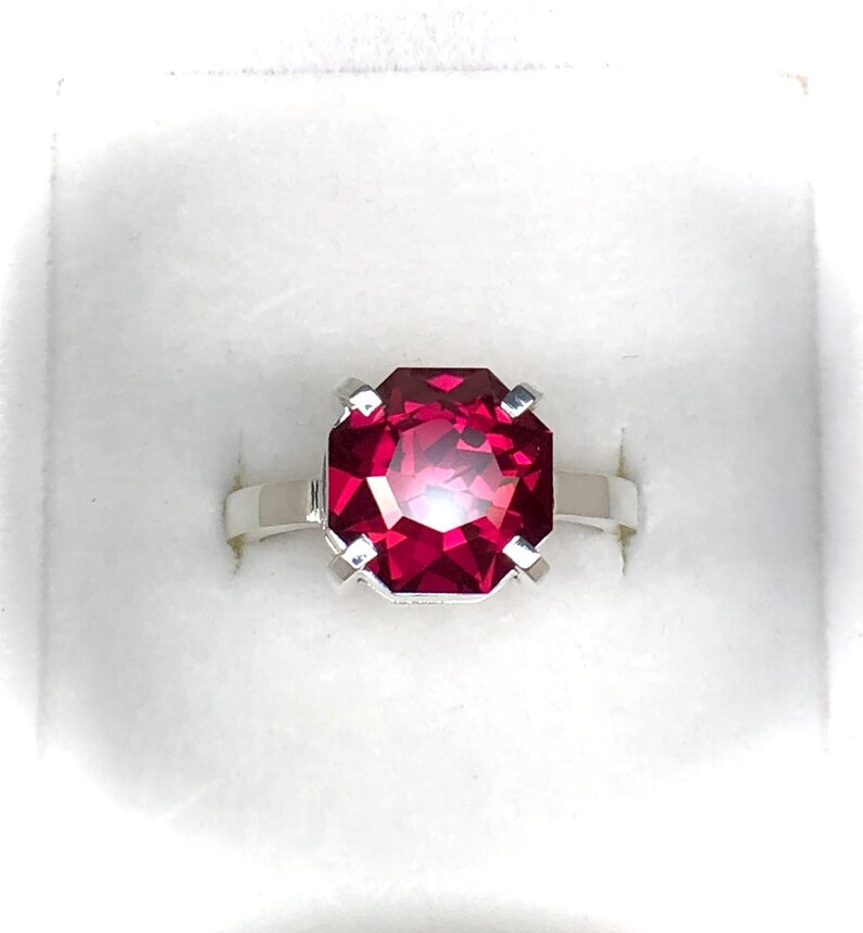May include: A silver ring featuring a large, faceted, rectangular-cut ruby gemstone. The ruby is a vibrant shade of red, and the ring is set in a simple, elegant design. The ring is displayed in a white box.