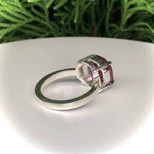 May include: A silver ring with a large, rectangular, ruby-colored gemstone. The gemstone is set in a geometric, open-work setting. The ring band is smooth and polished.