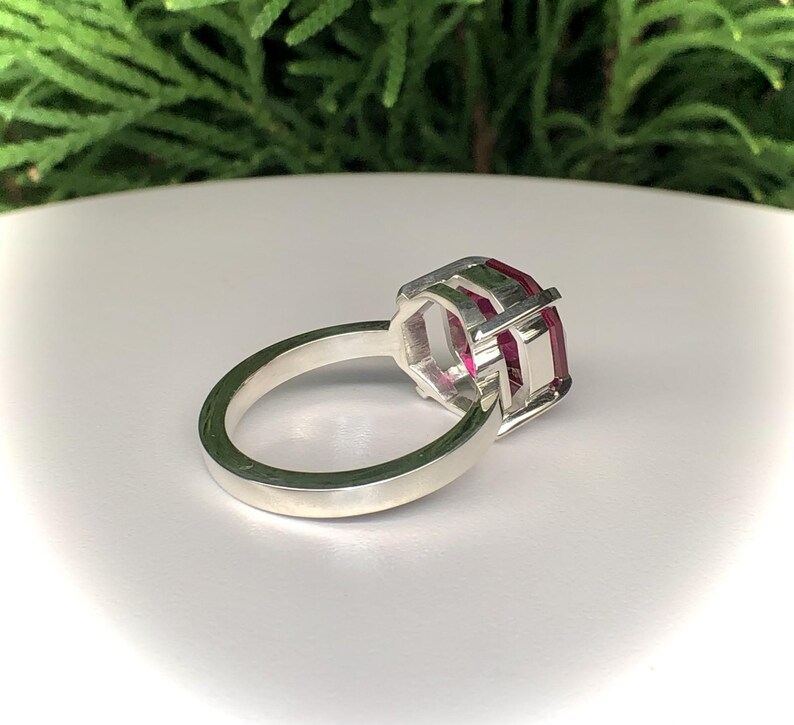 May include: A silver ring with a large, rectangular, ruby-colored gemstone. The gemstone is set in a geometric, open-work setting. The ring band is smooth and polished.