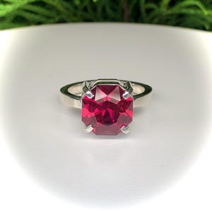 May include: A silver ring featuring a large, faceted, rectangular-cut, deep red gemstone. The gemstone is held in place by four prongs. The ring is set against a white background.