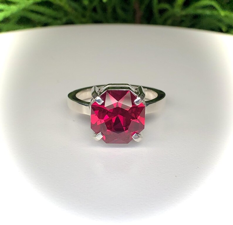 May include: A silver ring featuring a large, faceted, rectangular-cut, deep red gemstone. The gemstone is held in place by four prongs. The ring is set against a white background.