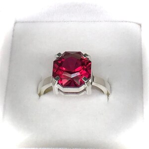 May include: A silver ring featuring a large, faceted, rectangular ruby gemstone. The ruby is a deep, vibrant red color, and the ring is set in a simple, elegant design. The ring is presented in a white box.
