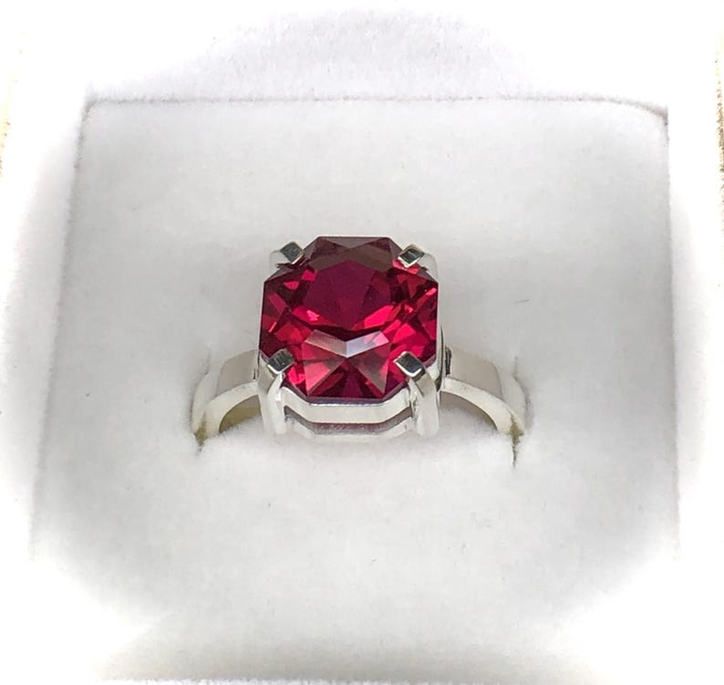 May include: A silver ring featuring a large, faceted, rectangular ruby gemstone. The ruby is a deep, vibrant red color, and the ring is set in a simple, elegant design. The ring is presented in a white box.