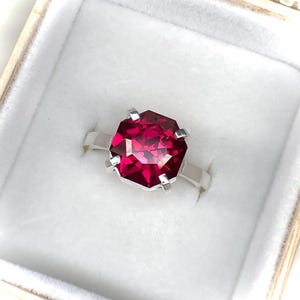 May include: A silver ring with a large, faceted, ruby-red gemstone. The ring is set in a simple, modern style, and is presented in a white jewelry box with a soft, velvety interior.